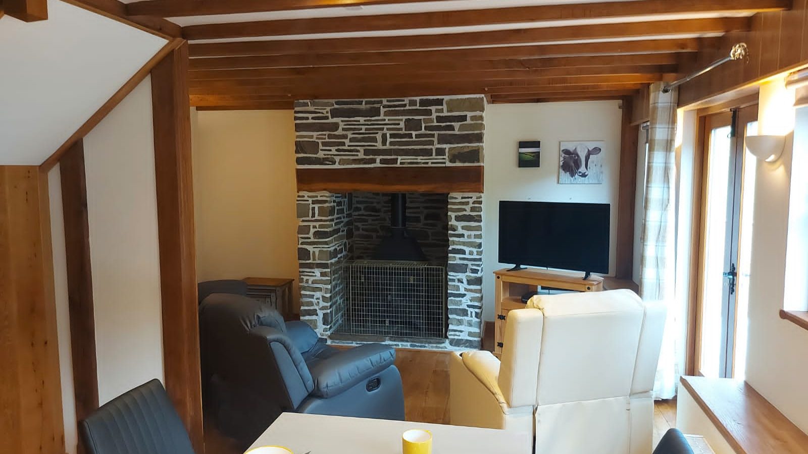 Cozy living room in Ploony, Wales, featuring exposed beams and a stone fireplace. Perfect for holidays, it includes a TV, comfortable armchairs, and a dining table set elegantly with yellow dinnerware.