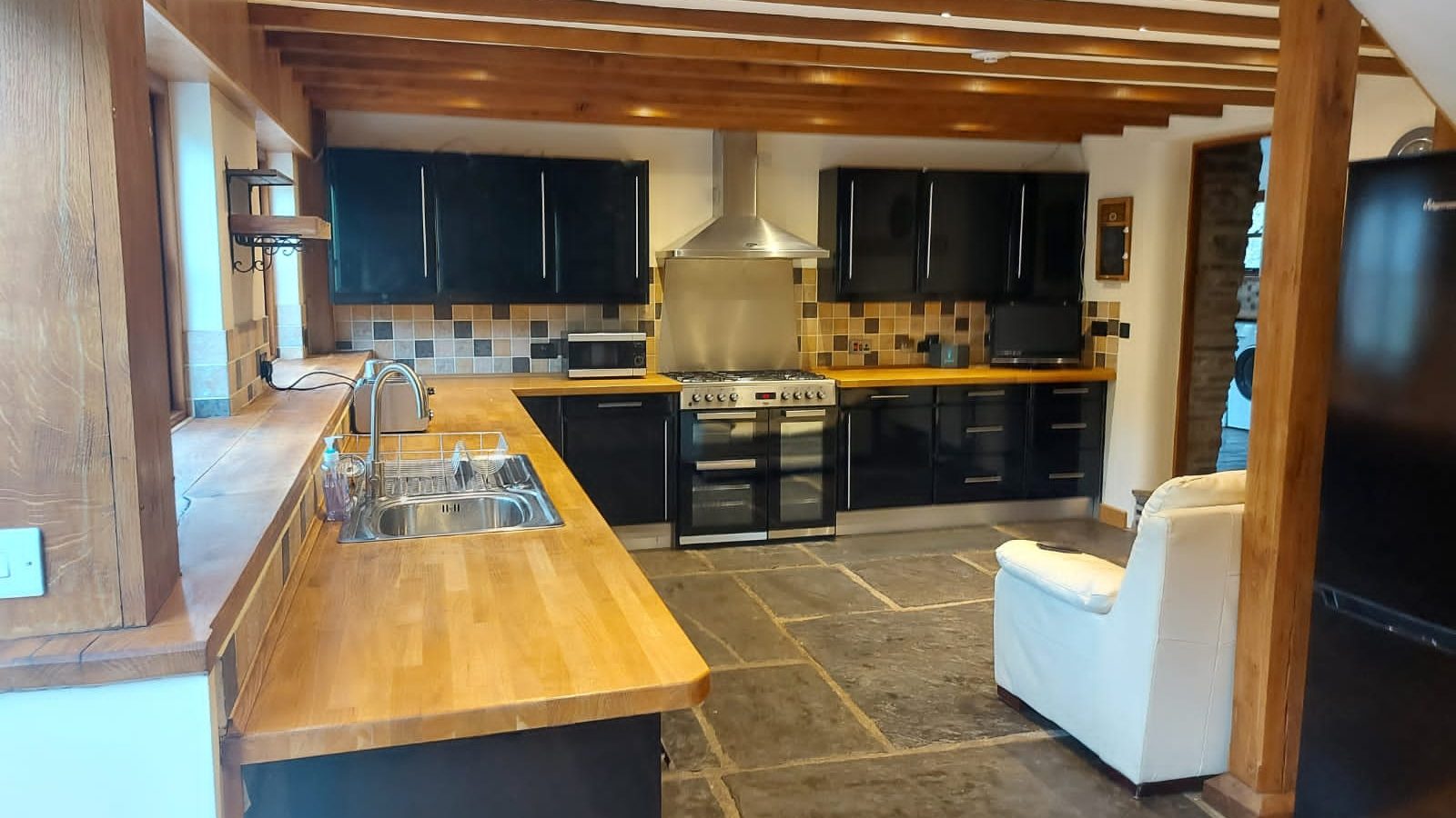 Modern kitchen with wooden countertops, tiled backsplash, and black cabinets. Features a double sink, oven, and cushioned chair—perfect for preparing festive holiday feasts inspired by the rustic charm of Wales.