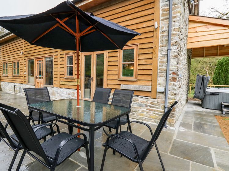 The outdoor patio, perfect for Ploony Holidays in Wales, features a black table and chairs under a large umbrella beside a wooden and stone house with expansive windows.