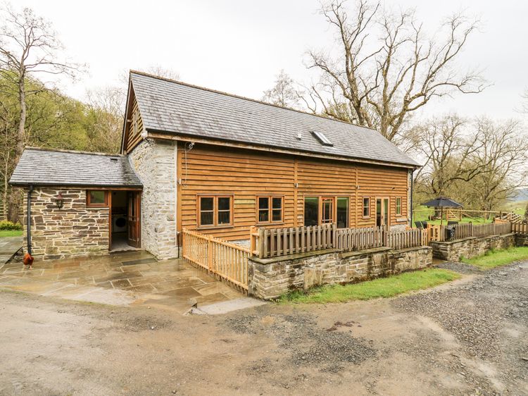 Nestled in the heart of Wales, this charming rustic wooden and stone house with a slate roof is surrounded by trees. Perfect for Ploony Holidays, it features a cozy patio with a table and umbrella, inviting you to relax in nature’s embrace.