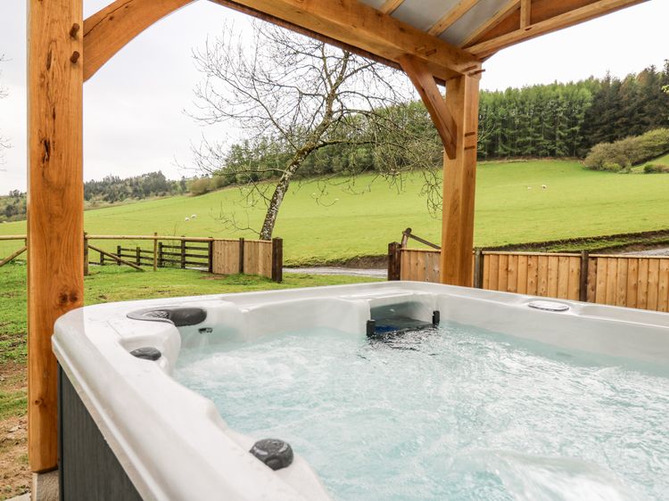 This outdoor hot tub under a wooden shelter offers a serene escape with stunning views of a grassy field and trees in the background. Experience the tranquility of Wales at Ploony Holidays, where nature and relaxation seamlessly blend together.