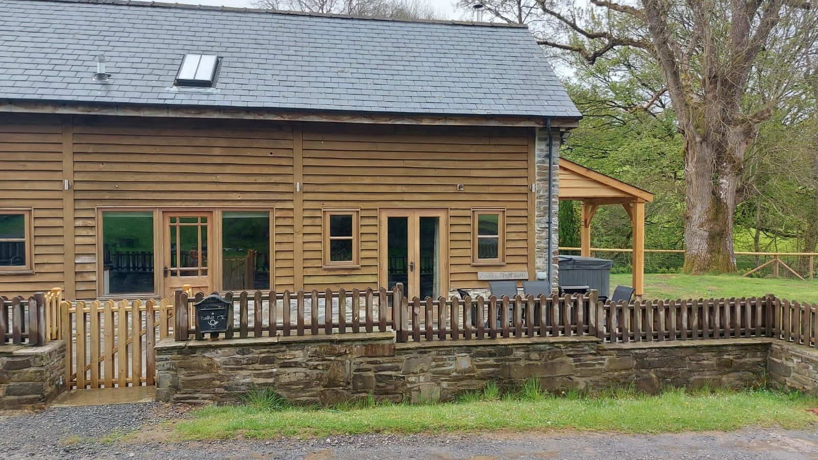 A charming wooden building with a slate roof and fenced yard, surrounded by lush trees, rests on a gravel path. Perfect for a serene escape, this gem embodies the tranquility of Wales and is ideal for those seeking Ploony Holidays.