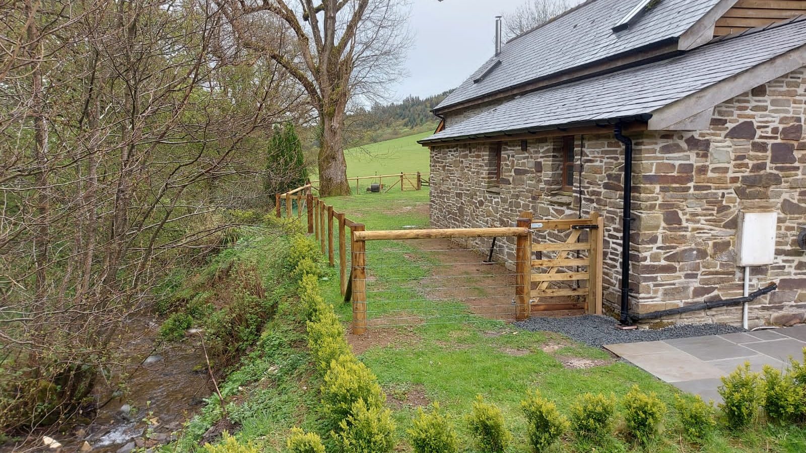 Nestled in the serene landscapes of Wales, this rural stone house with a wooden fence sits beside a gentle stream, surrounded by lush trees and grassy fields—an idyllic retreat offering the timeless charm of Ploony Holidays.