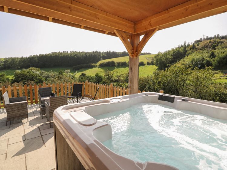 Experience Ploony Holidays with an outdoor hot tub under a wooden pergola, boasting breathtaking views of Wales' green hills and trees. Nearby, a cozy seating area with chairs and table invites relaxation amidst nature's splendor.