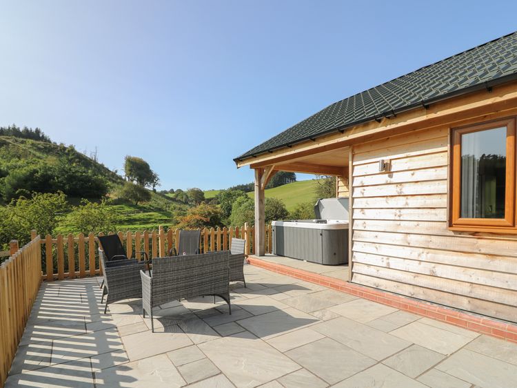 Experience a Ploony holiday in this charming wooden cabin with a patio featuring wicker chairs and a hot tub, overlooking the scenic hills and lush trees of Wales.