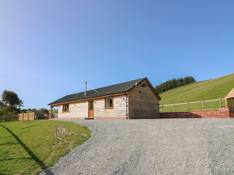 Nestled on a gravel driveway, the wooden cabin boasts a sloped roof and is surrounded by the lush green hills of Wales under a clear blue sky, promising an idyllic retreat perfect for Ploony Holidays.