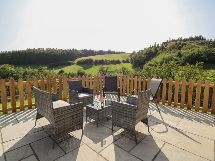 The patio features wicker furniture and a glass table, perfect for enjoying the lush green landscape of Wales. Surrounded by a wooden fence, it's an idyllic spot that captures the essence of holidays at Ploony.