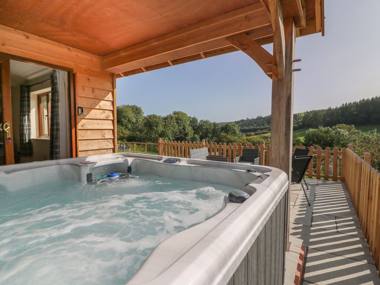 Enjoy a relaxing soak in the outdoor hot tub on a wooden deck with cozy patio furniture, all while taking in the scenic view of trees and hills. Perfect for your getaway with Ploony Holidays in beautiful Wales.