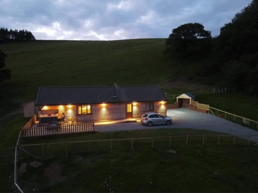 A single-story house with lights on sits serenely in rural Wales, a car parked outside beneath the cloudy evening sky, offering the perfect retreat for those seeking Ploony Holidays.