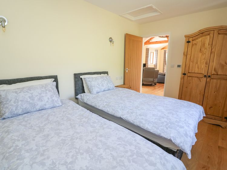 Twin beds with floral bedding adorn a cozy bedroom featuring a wooden door and wardrobe, offering a perfect retreat for Ploony Holidays. A glimpse of the inviting living area is visible through the door, capturing the charm of this idyllic spot in Wales.