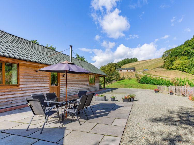 Enjoy a relaxing holiday in Ploony, with a charming patio beneath an umbrella offering a peaceful retreat adjacent to a wooden building, all set against the serene rural landscape of Wales.