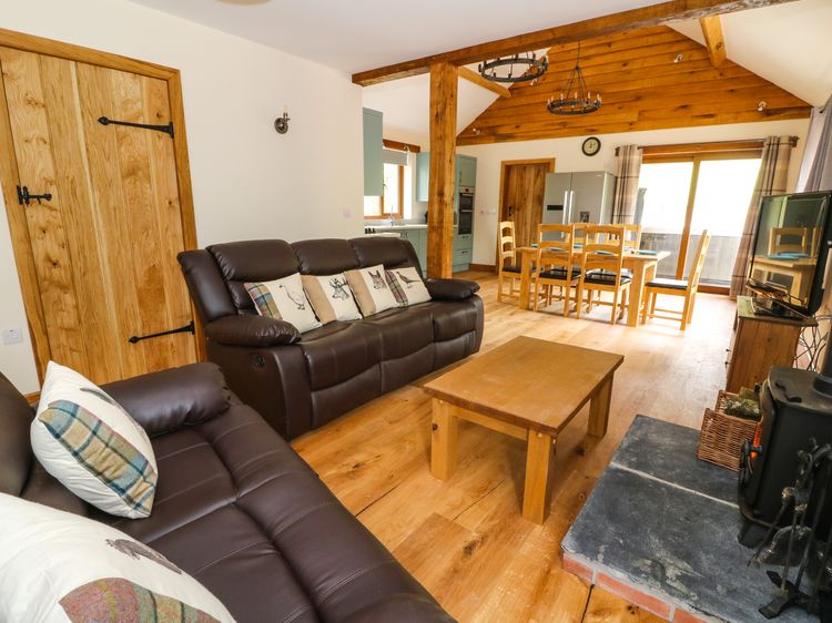 A cozy living room perfect for a Ploony Holidays retreat in Wales, featuring leather sofas, a wooden coffee table, dining area, and a fireplace. Exposed beams add rustic charm.