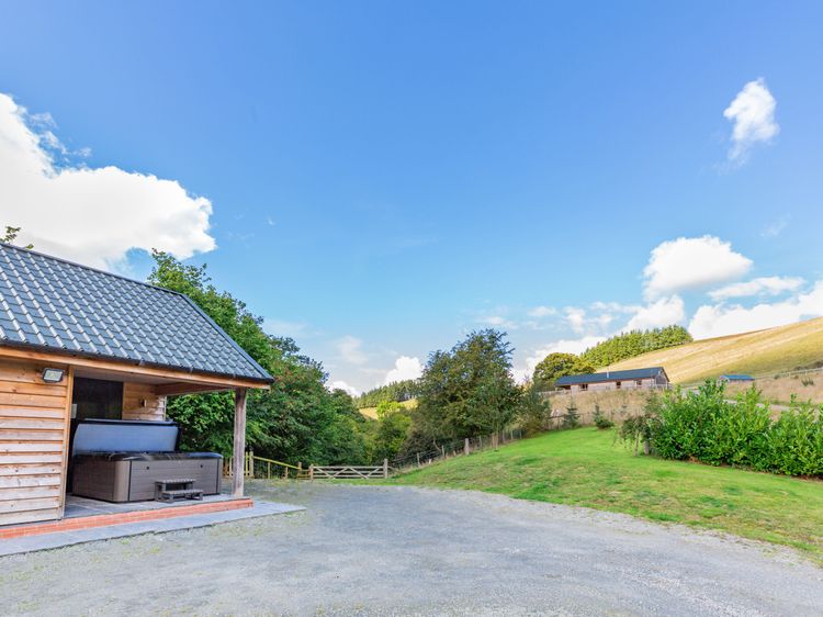 Nestled in the serene landscape of Wales, this Ploony Holidays wooden cabin features a covered hot tub that overlooks a grassy field and distant hills under a blue sky with clouds.