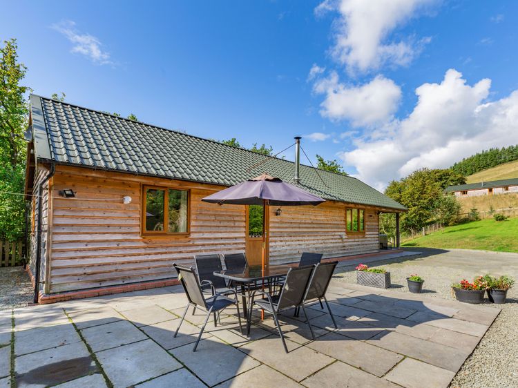 Nestled in the serene landscapes of Wales, this charming wooden house features a green roof and offers a patio set with six chairs under an umbrella. The gravel driveway in the foreground invites you to begin your Ploony Holidays adventure.