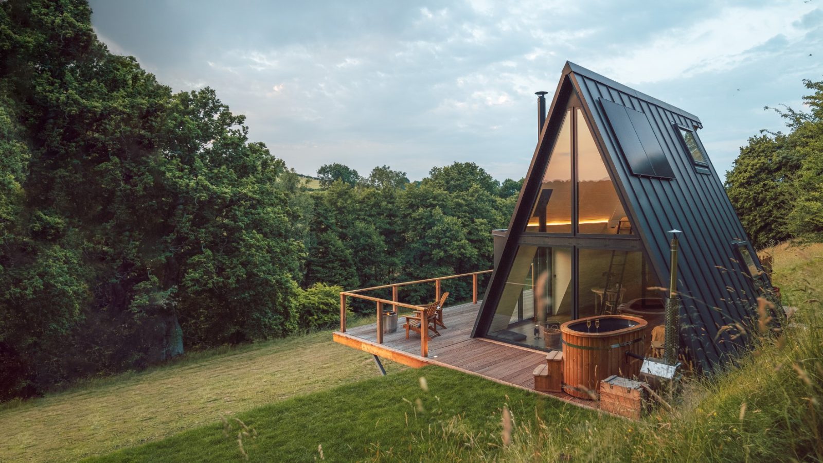 The Fold, an A-frame cabin with large windows and a wooden deck featuring a hot tub, is set on a hillside surrounded by lush greenery.