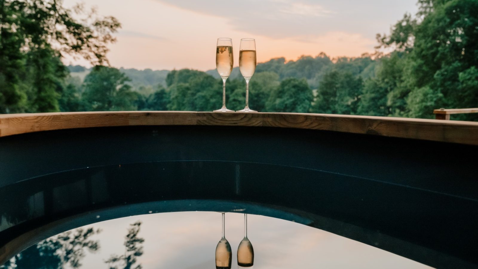 Two champagne glasses rest on a wooden railing, framed by a scenic view of trees and a vibrant sunset in the background.