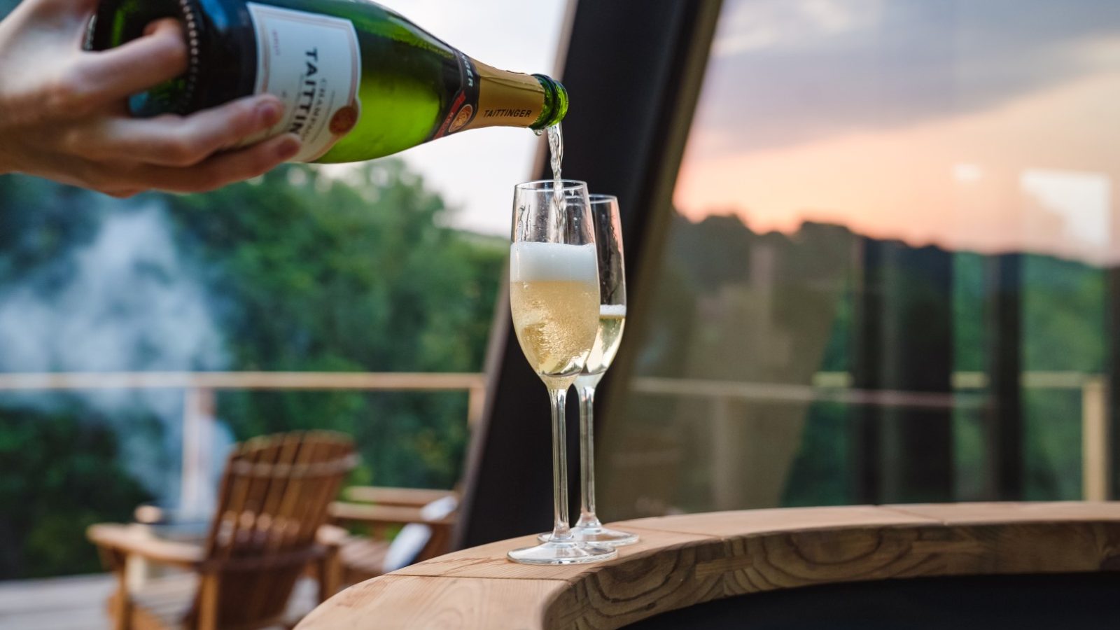 A person pours champagne into a flute glass on a table at The Fold, with a scenic outdoor view in the background.