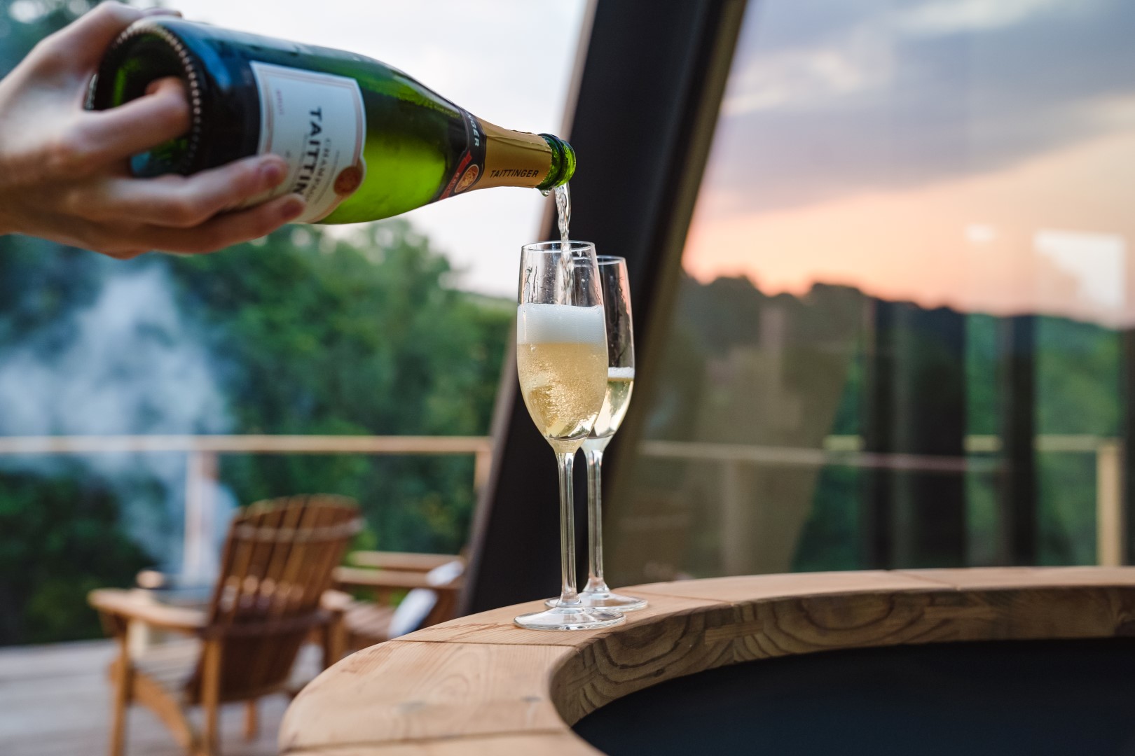 A person pours champagne into a flute glass on a table at The Fold, with a scenic outdoor view in the background.