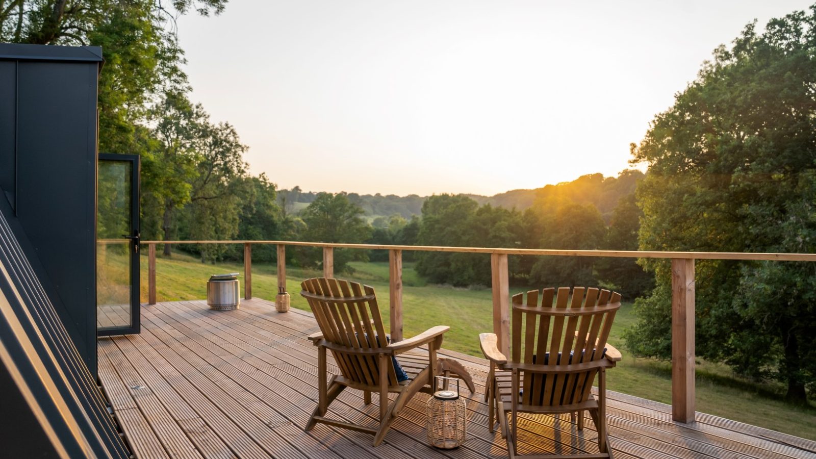 Two wooden chairs on the deck of The A Frame overlook a green field with trees, under a clear sky during sunset.