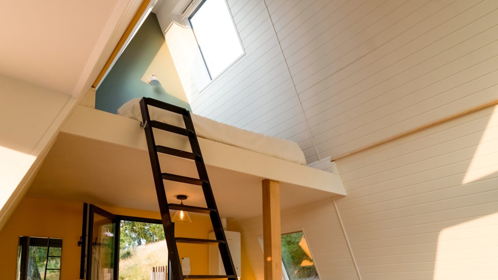 A small loft bed inside an A Frame room with a sloped ceiling, accessed by a wooden ladder, over a vibrant yellow-walled lower area.