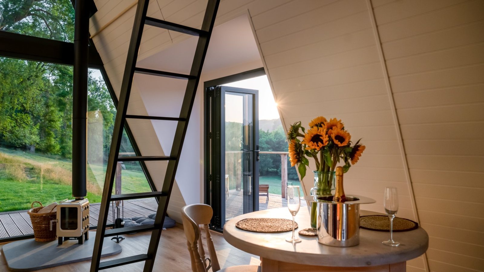 Cozy A-frame cabin interior at The Fold with a round table, sunflowers in a vase, and a view of greenery through an open glass door.