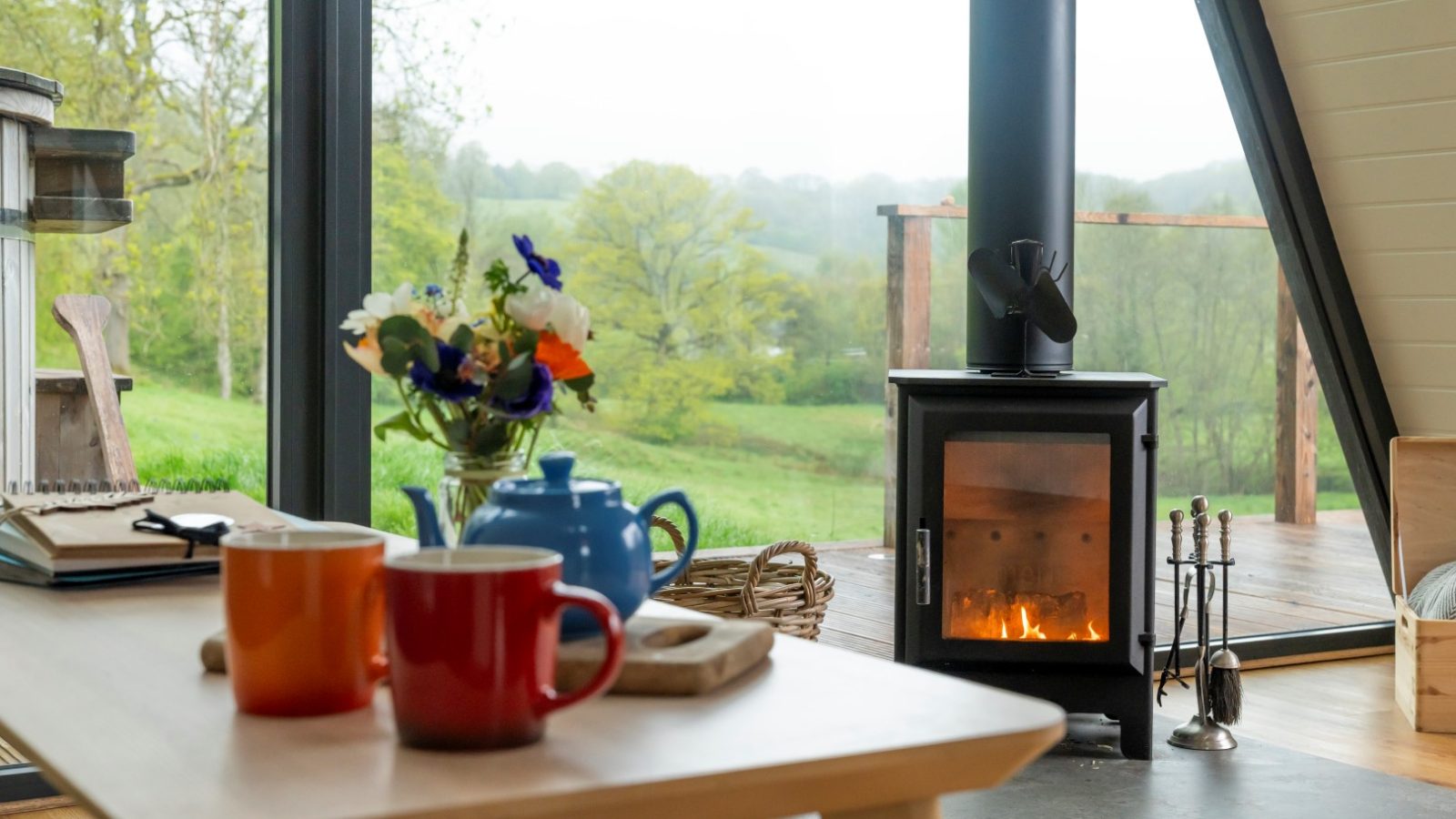 A cozy A Frame living room with a wood-burning stove, two mugs, a teapot, and a vase of flowers on a table, overlooking a green landscape.