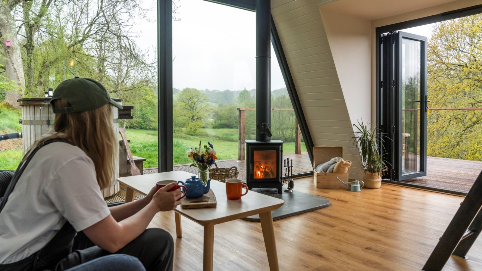 A person sits at a table with mugs and a teapot, gazing out at the scenic view from a cozy A-Frame cabin, its warmth enhanced by large windows and a crackling wood stove.