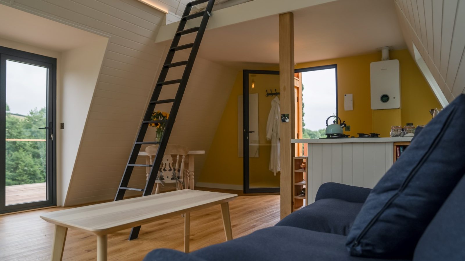 Loft-style interior reminiscent of The Fold, featuring a ladder to a cozy bed, a couch, wooden table, and a small kitchen area. Enjoying an outdoor view through the windows.