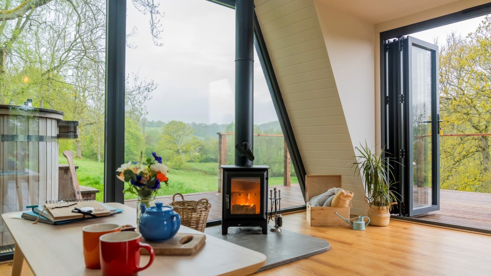 Bright A-frame room with large windows, wood stove, open doors, and a view of greenery. A teapot and mugs sit on a wooden table.