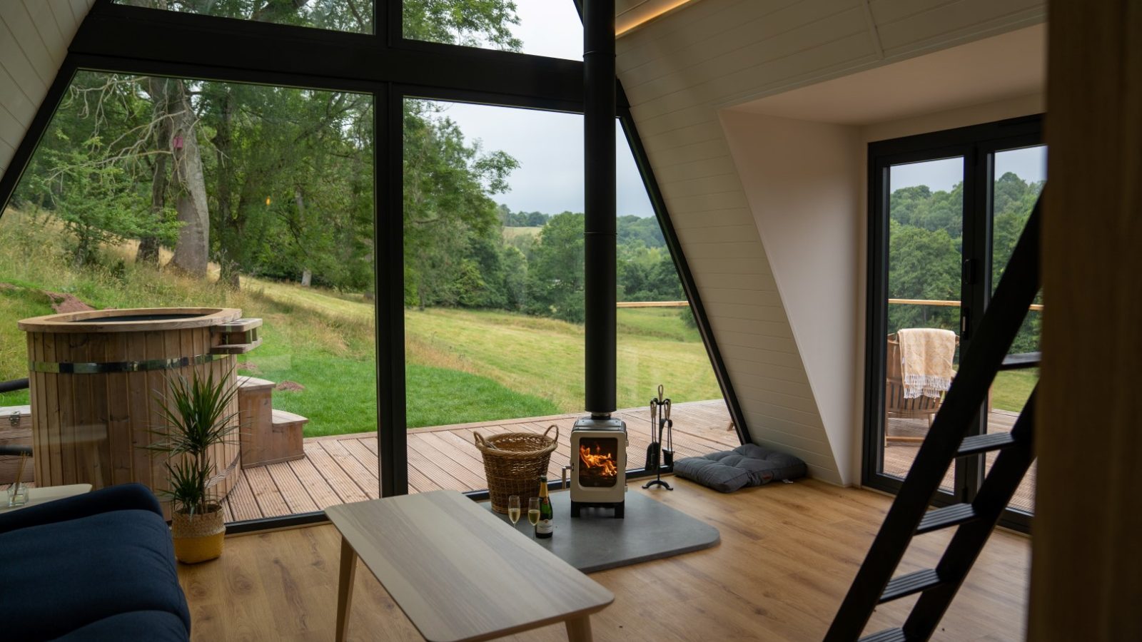 The Fold's A-frame cabin interior boasts large windows, wooden floors, and a cozy wood stove. Enjoy the serene view of greenery and unwind in the inviting hot tub outside.