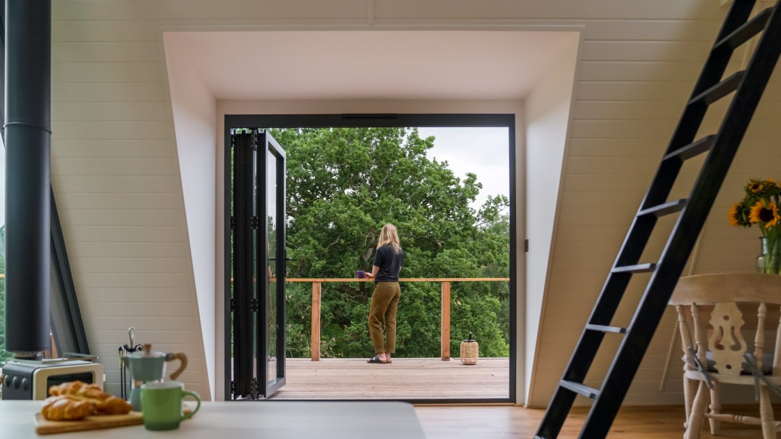 A woman stands on a wooden deck designed in an A Frame style, overlooking lush green trees, viewed from inside a cozy room with a ladder and breakfast items.