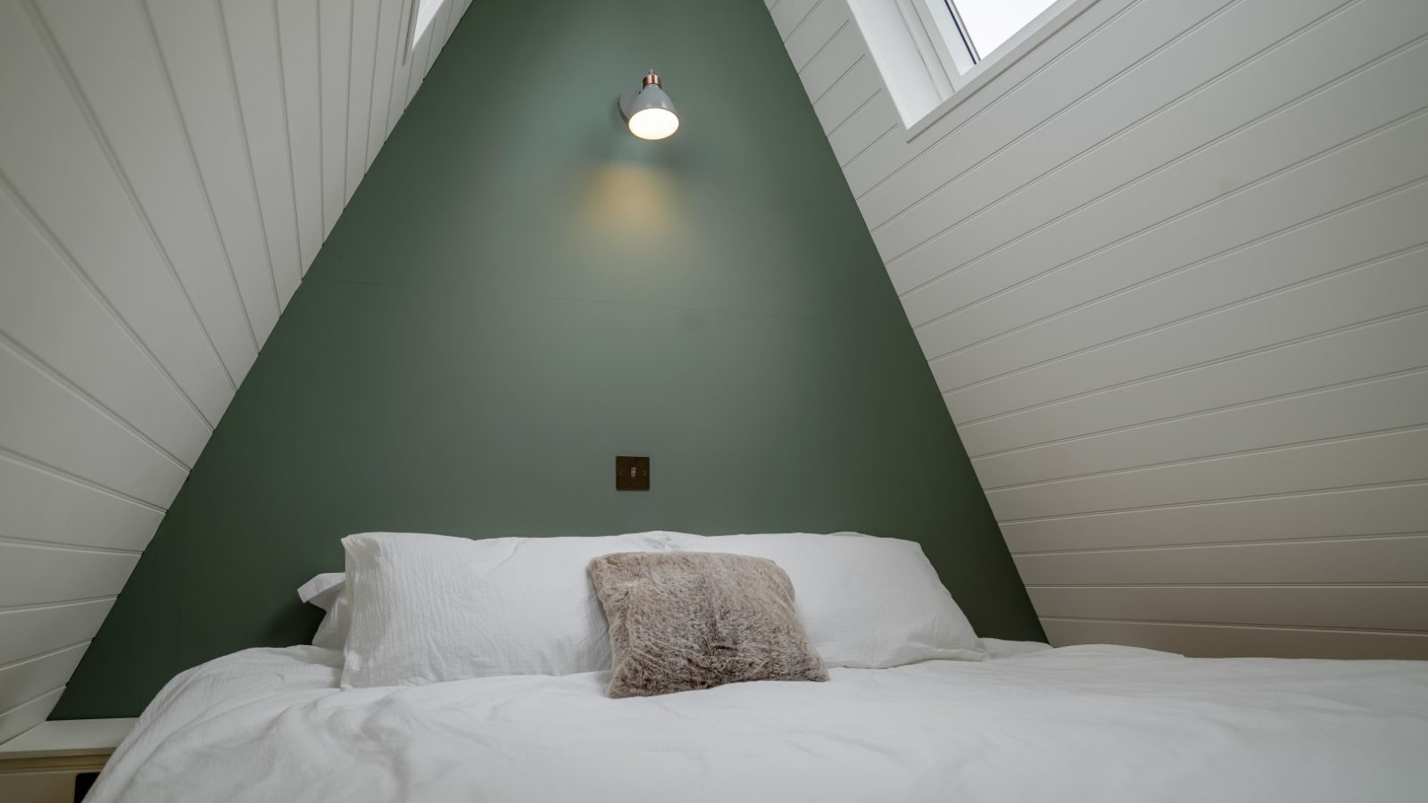 A cozy bedroom in The Fold with a green accent wall, featuring an inviting bed with white linens, a small pillow, and an elegantly wall-mounted light fixture.