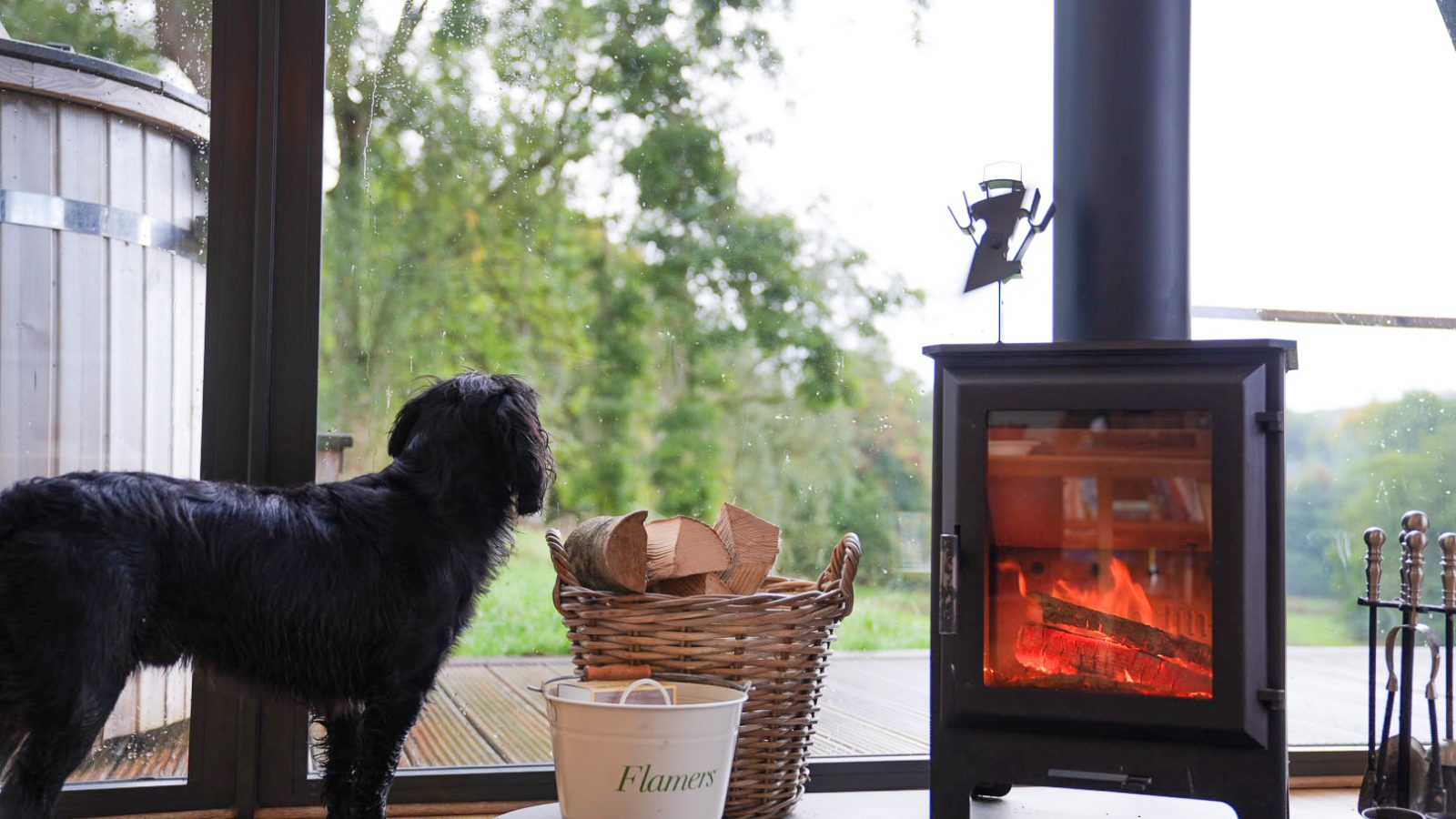 A black dog looks out an A-frame window beside a lit wood stove, with a basket of firewood and a bucket labeled 