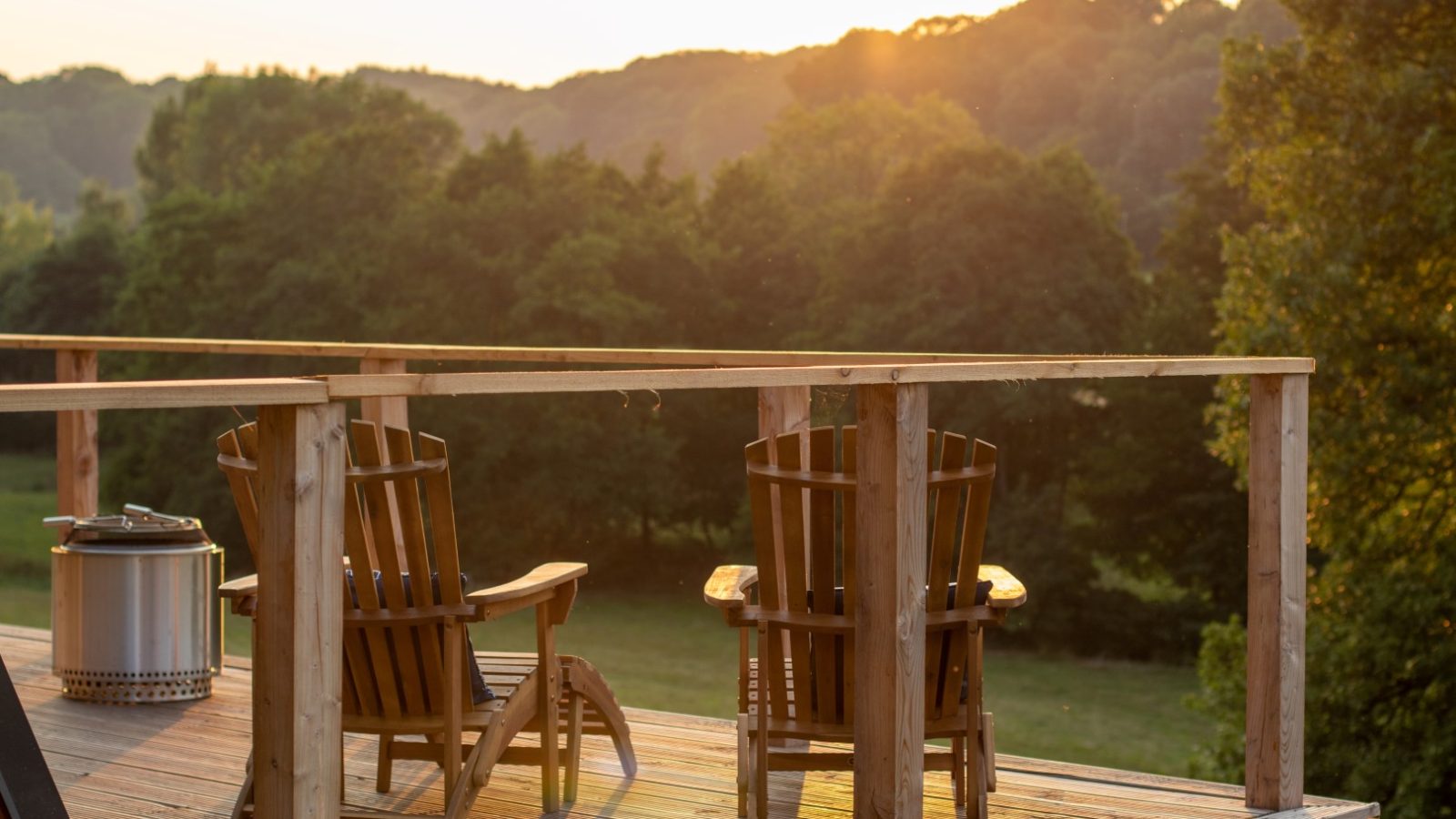 Two wooden chairs on a deck, reminiscent of an A Frame design, overlook a lush green landscape at sunset, with trees and hills stretching into the horizon.