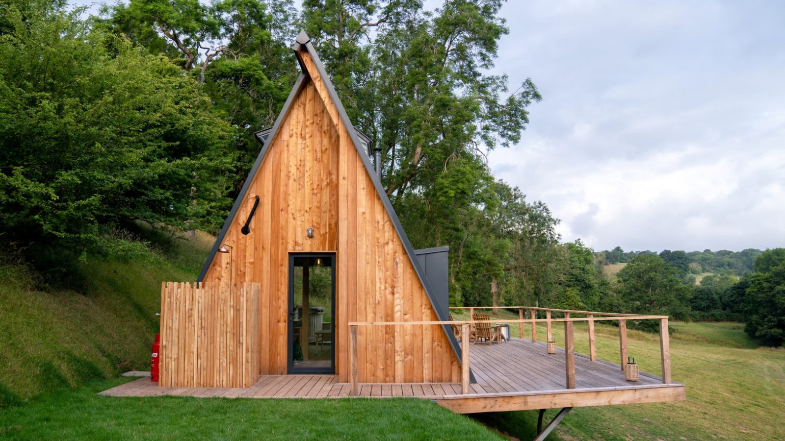 A small, A-frame wooden cabin with a sloped roof sits on a grassy hill, surrounded by trees, overlooking a field.