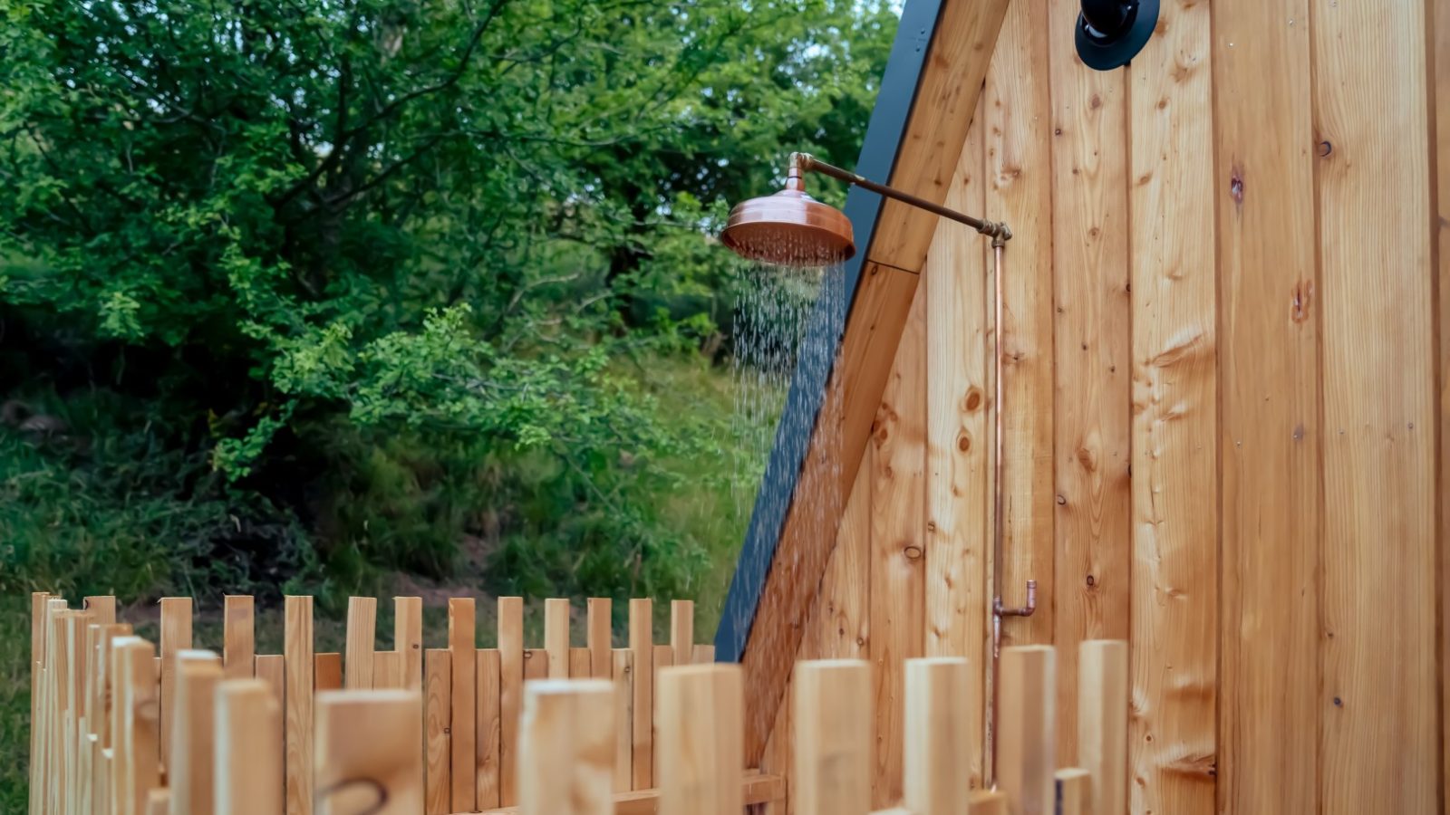 An outdoor shower at The Fold, featuring a wooden fence and wall, nestled amid lush greenery. Water gracefully cascades from a copper showerhead.