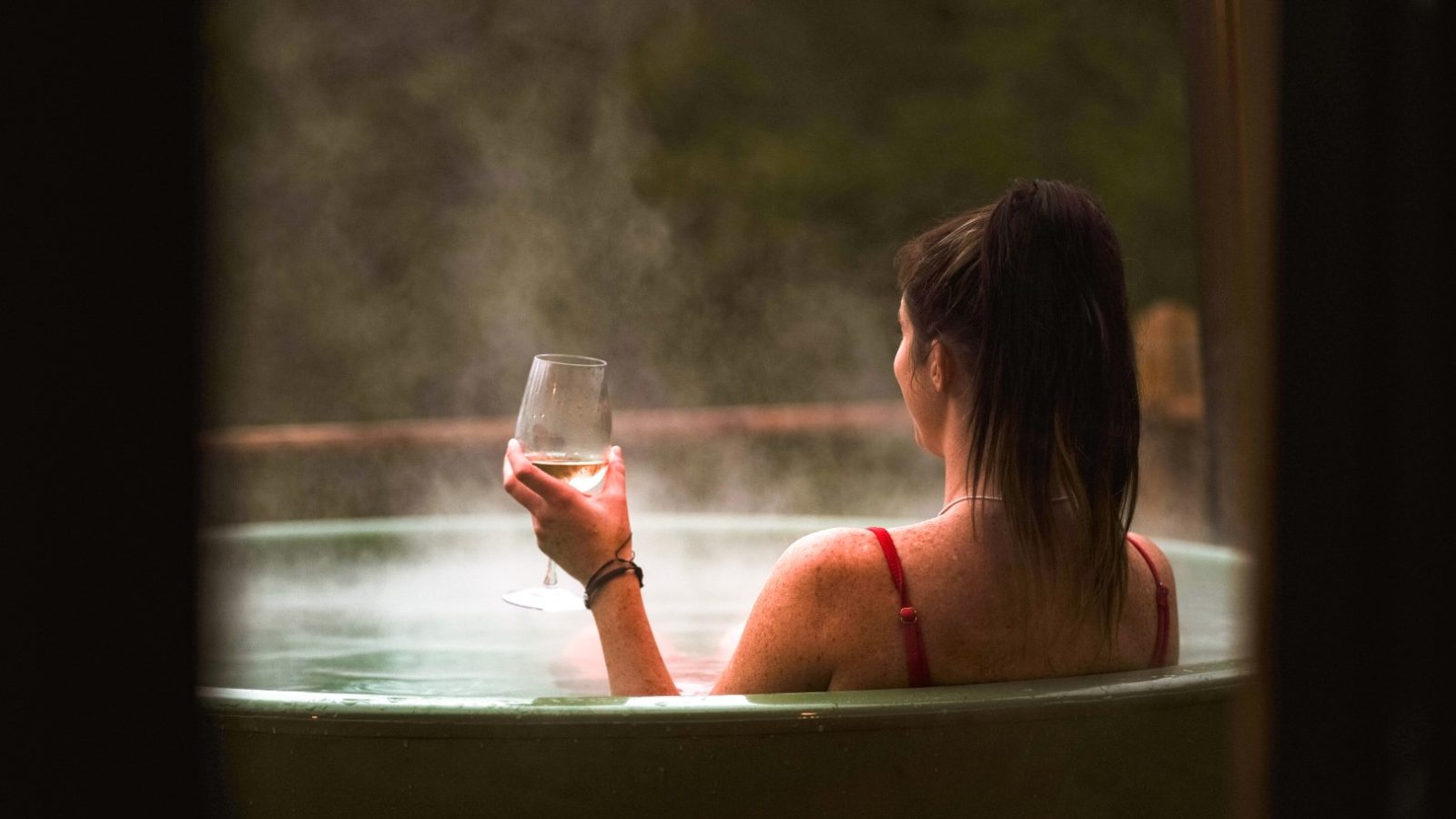 In the serene outdoor setting of The Fold, a person relaxes in a hot tub, steam rising around them as they savor the moment with a wine glass in hand. It's the perfect Scandi cabin escape.