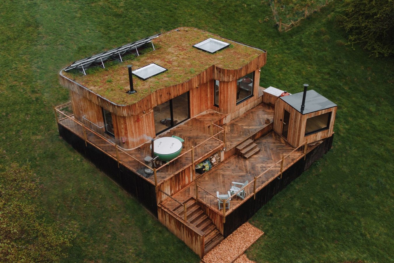 Aerial view of The Fold, a modern Scandi cabin with a grass-covered roof, hot tub on the deck, and chairs, all surrounded by lush greenery.