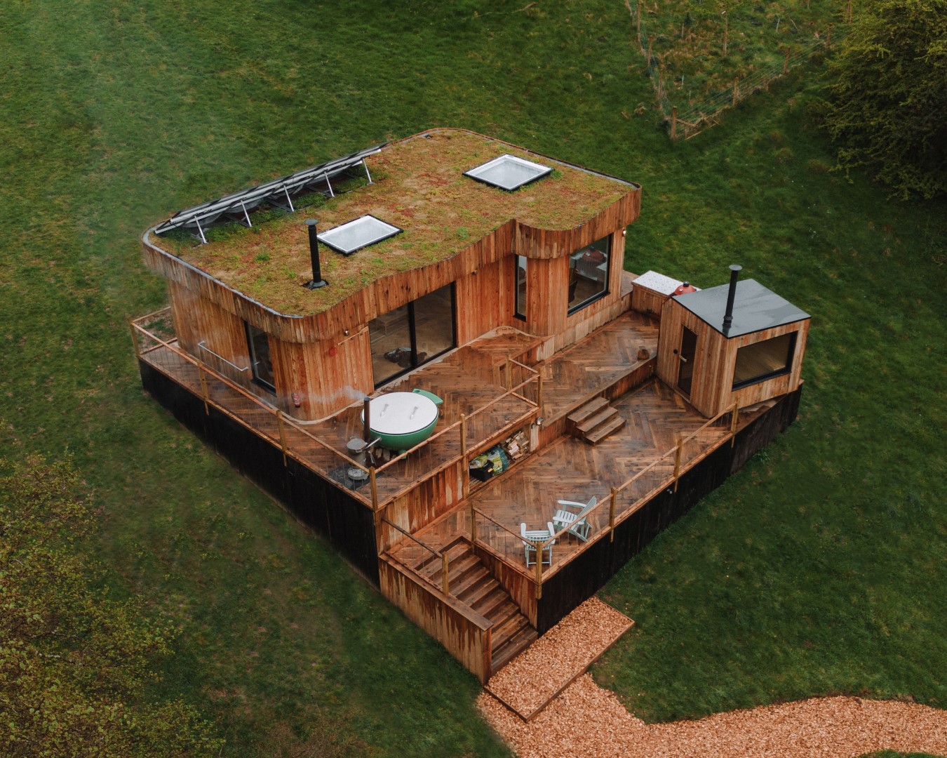 Aerial view of The Fold, a modern Scandi cabin with a grass-covered roof, hot tub on the deck, and chairs, all surrounded by lush greenery.