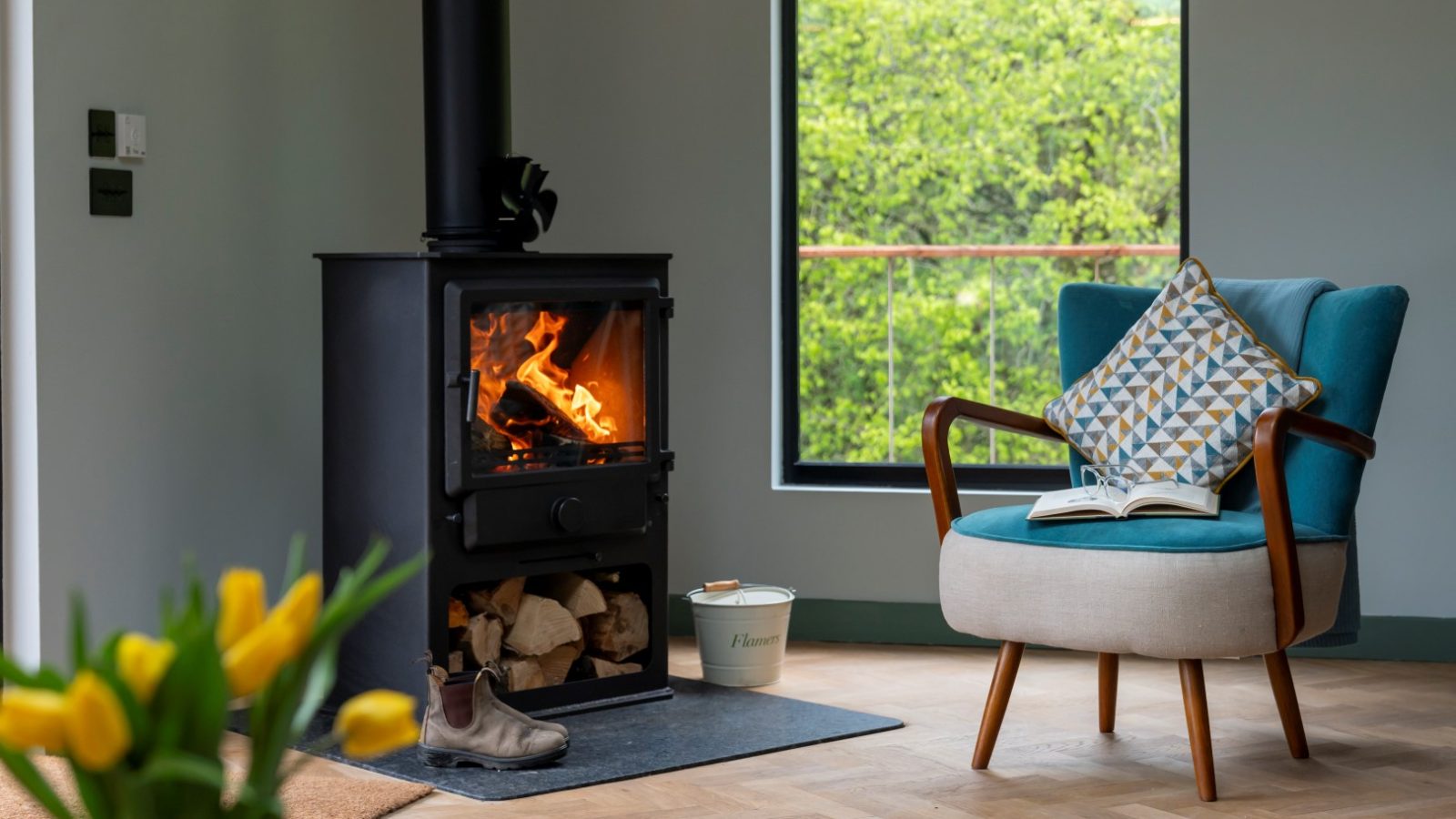 In this Scandi Cabin, a cozy room features a lit wood stove, a blue armchair with a pillow, an open book, and a vase of yellow tulips by the window, offering views of lush greenery.