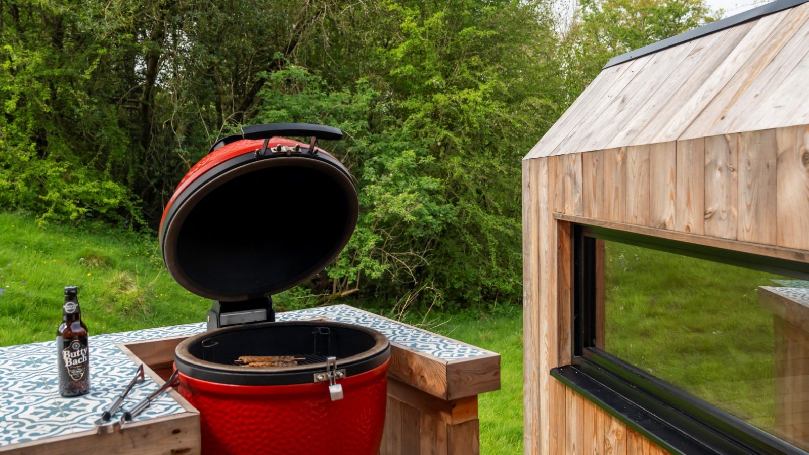 A red BBQ grill sits on a tiled patio next to a beer bottle and tongs, framed by lush trees and the rustic charm of The Fold's wooden structure.