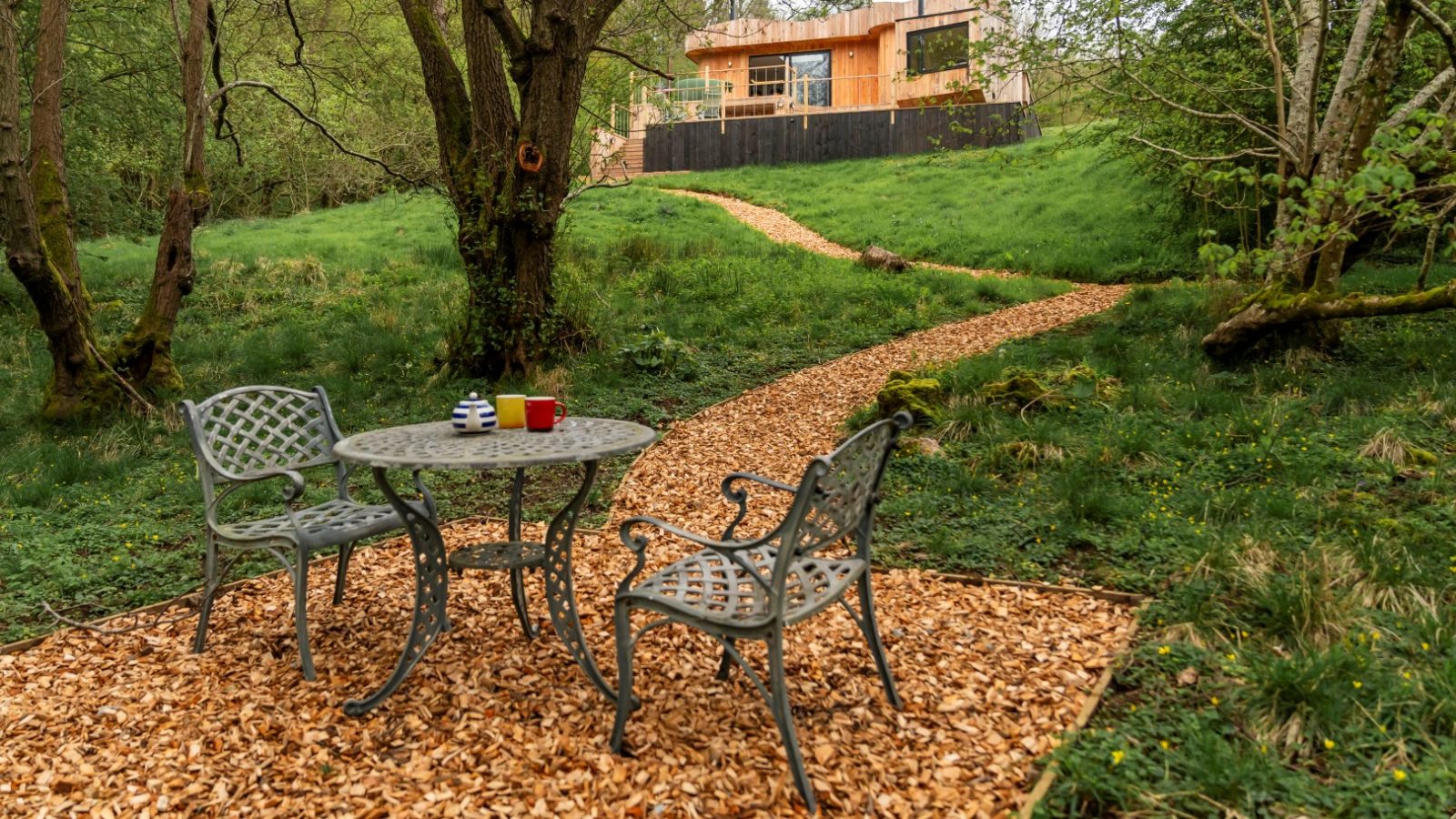 A pathway leads to The Fold, a modern house in the woods. In the foreground, a metal patio set is nestled amid trees and lush greenery, offering a serene retreat reminiscent of a Scandi Cabin.