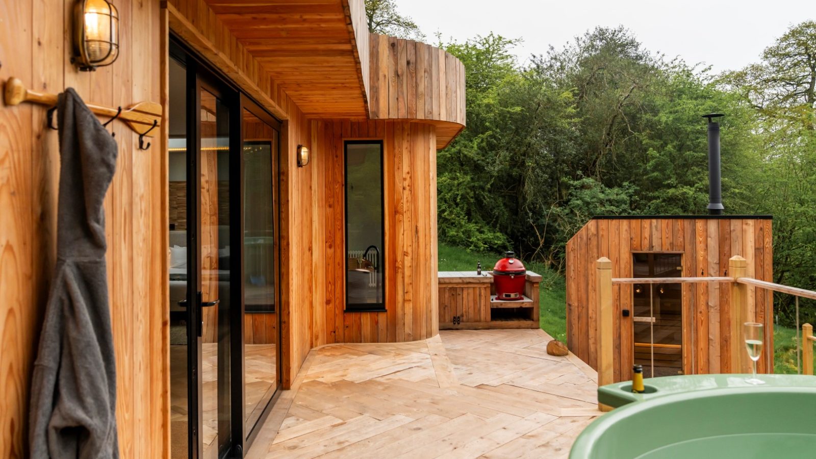 Nestled in the serene backdrop of towering trees, The Fold boasts a wooden deck with glass doors leading to a round green hot tub. This Scandi Cabin's charm is enhanced by its vivid red barbecue, making it the perfect retreat for relaxation and outdoor dining.