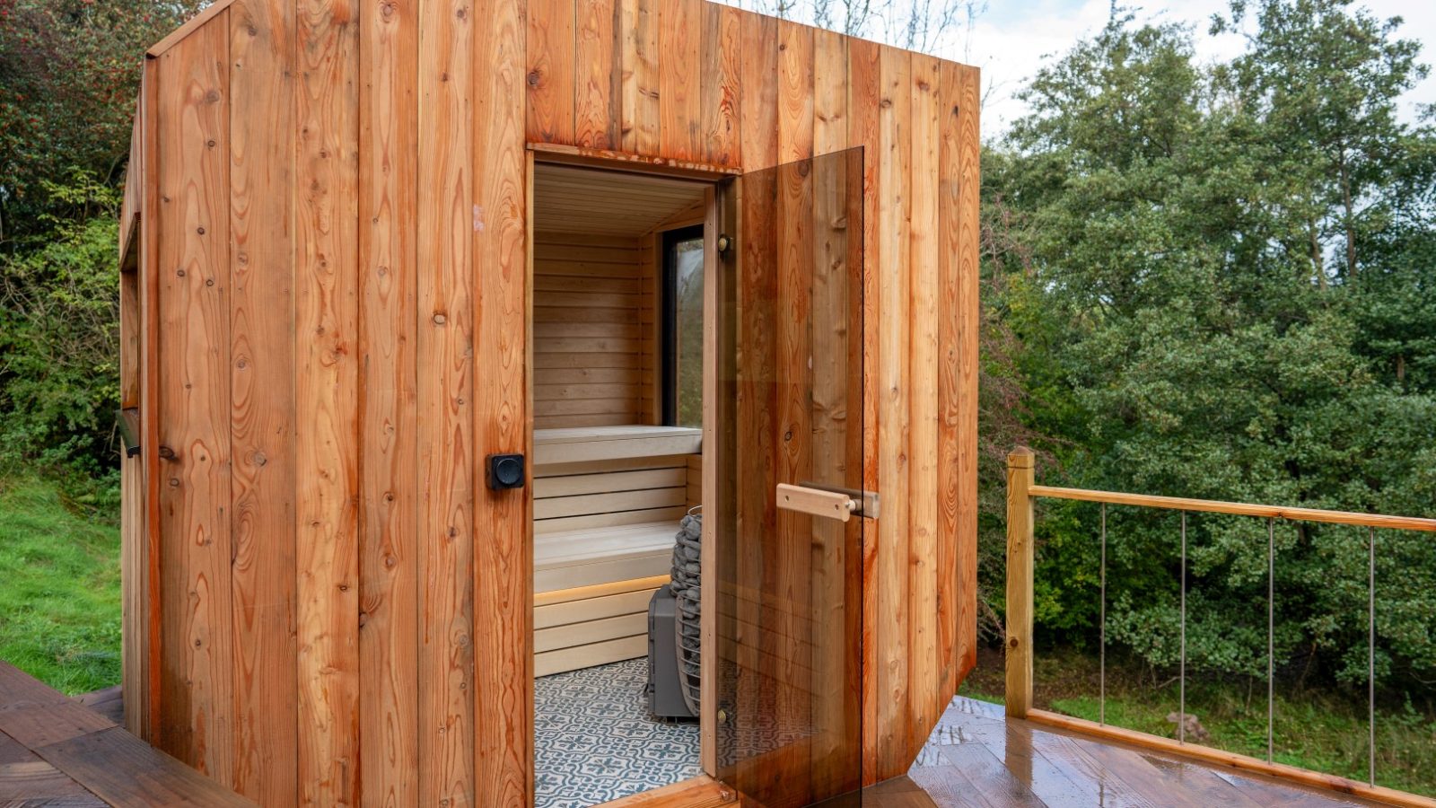 A wooden outdoor sauna with an open glass door, revealing cozy seating inside, resembles a charming Scandi Cabin. It's enveloped by lush trees and vibrant greenery.