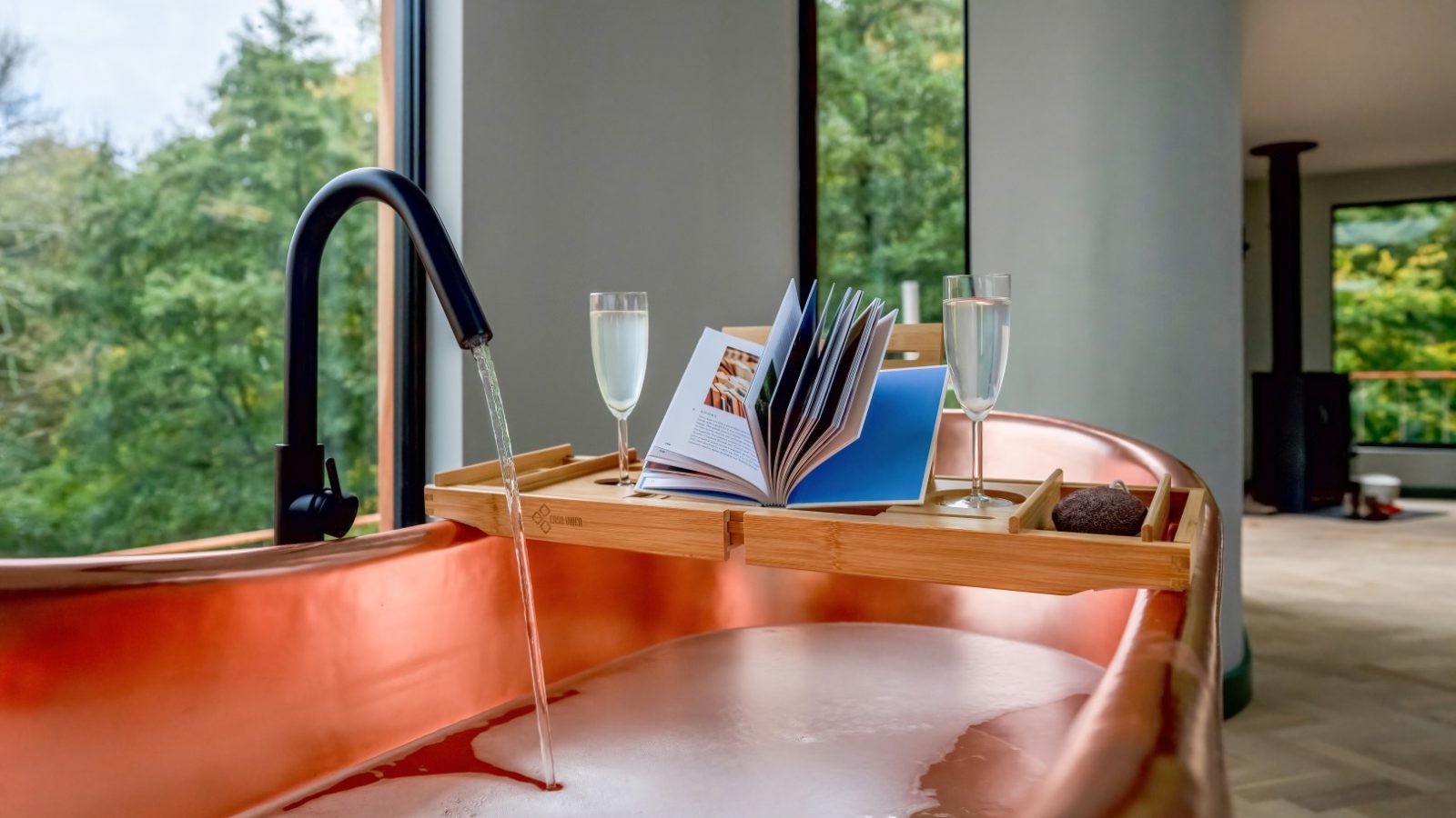 In the serene setting of a Scandi Cabin, a copper bathtub with running water invites relaxation. A wooden tray holds an open book, two glasses, and a candle, framed by large windows that offer an enchanting forest view.