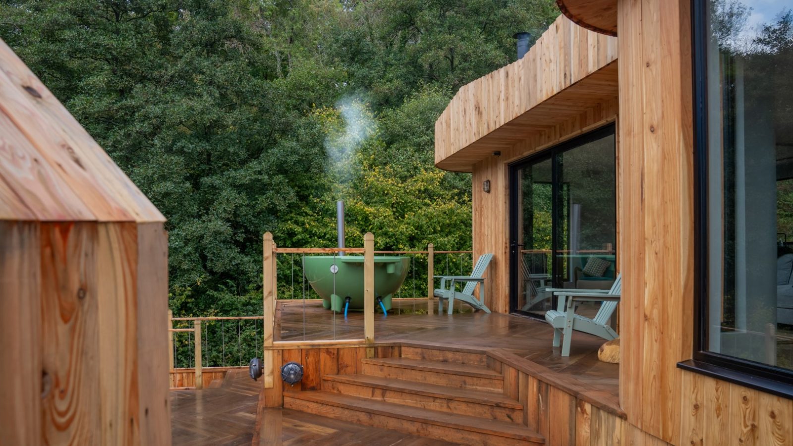 A wooden deck with steps leads to a green hot tub and chairs, enveloped by lush trees and set against the backdrop of a modern wooden building. This serene hideaway is reminiscent of a Scandi cabin, offering a tranquil oasis amid nature's embrace.