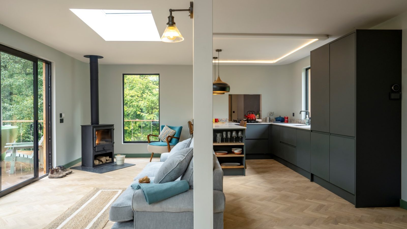 The Scandi Cabin features an open-plan living room and kitchen with a wood stove, grey sofa, dark cabinets, and large windows overlooking greenery.