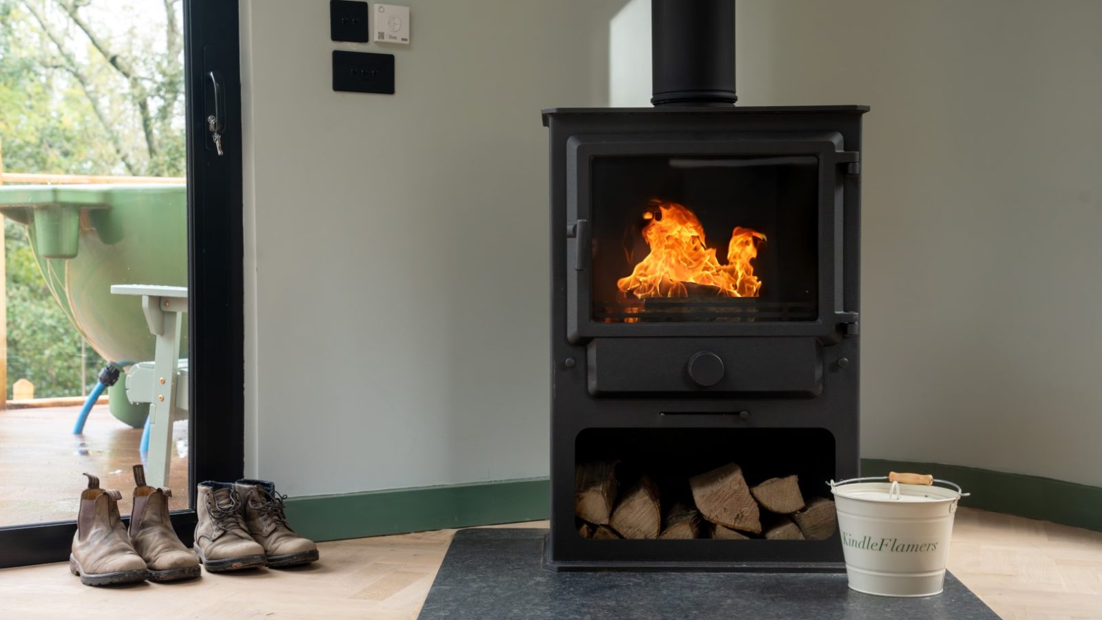 Wood-burning stove with visible flames, firewood stored below, and a bucket labeled 