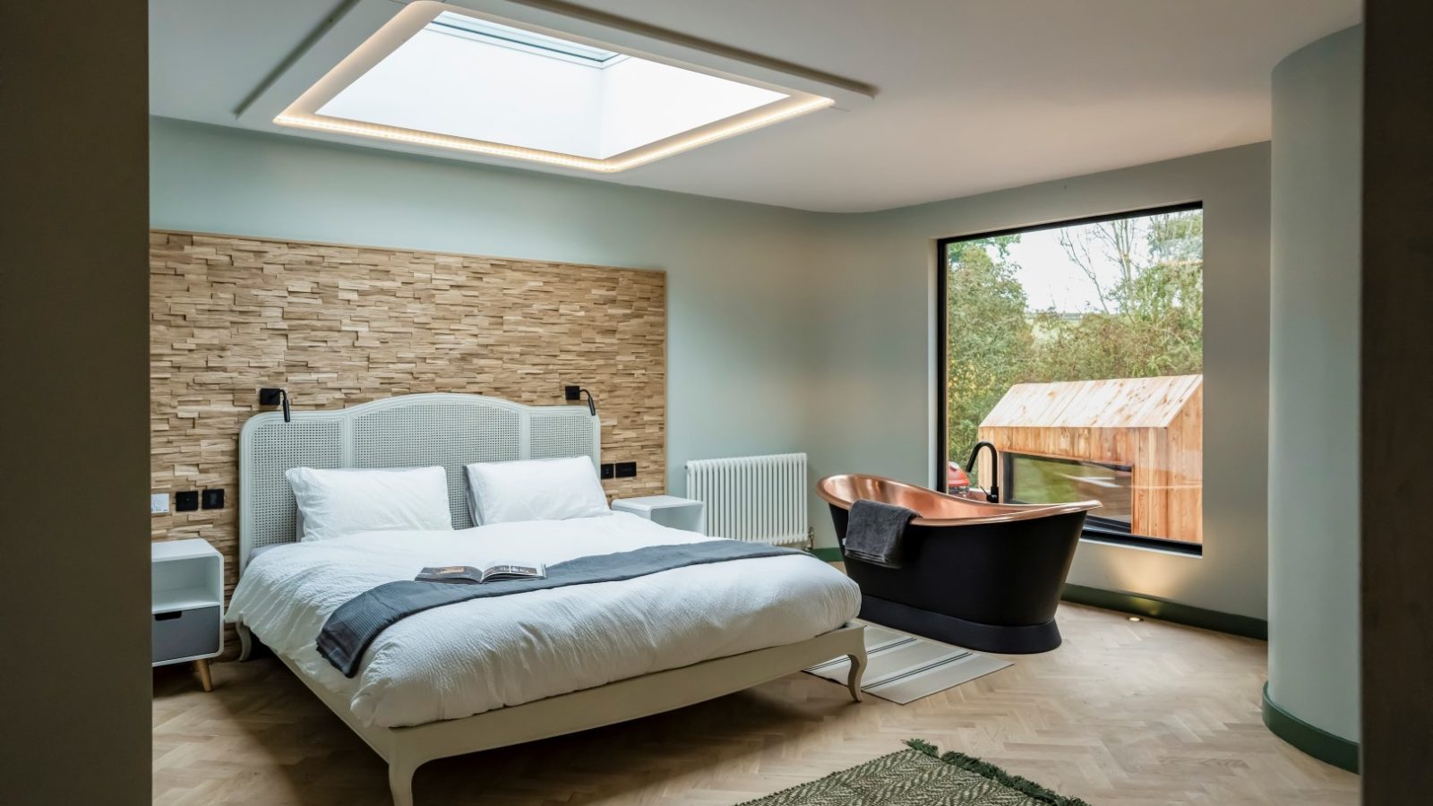 The Fold: a modern bedroom featuring a skylight, Scandi Cabin-inspired wooden accent wall, cozy bed, sleek black freestanding bathtub, and large window overlooking the garden.