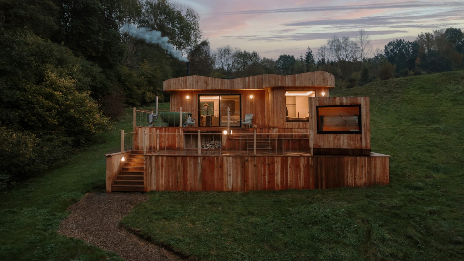 Scandi Cabin nestled on a grassy hill, featuring large windows and a deck perfect for sunset views, with trees forming a serene backdrop.